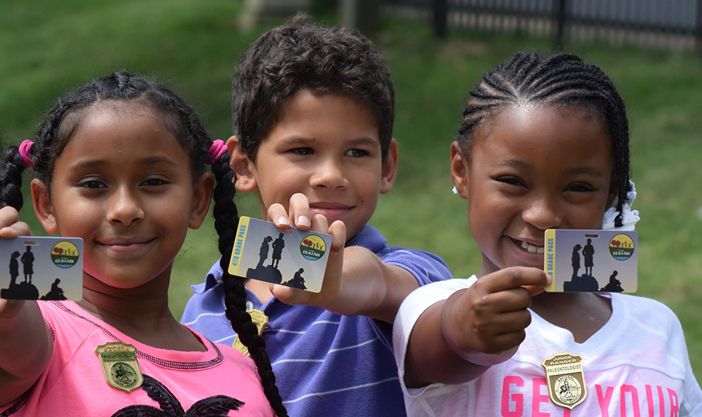 Three smiling children are holding up their free National Parks pass to the camera.